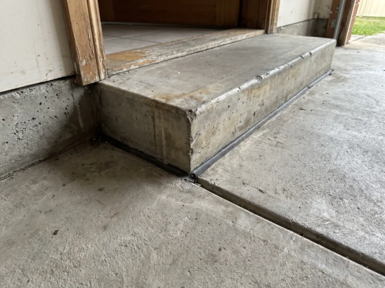 Concrete garage floor slab meeting a concrete step and door threshold, with a joint along the base of the step